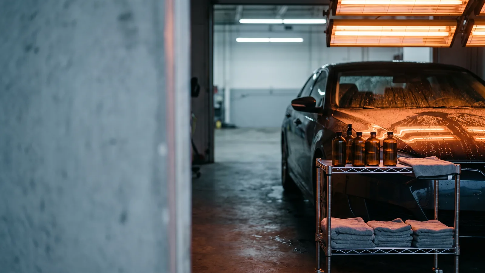 A dark sedan under curing lamps, half of its hood beading water, towels and plain bottles on a cart.