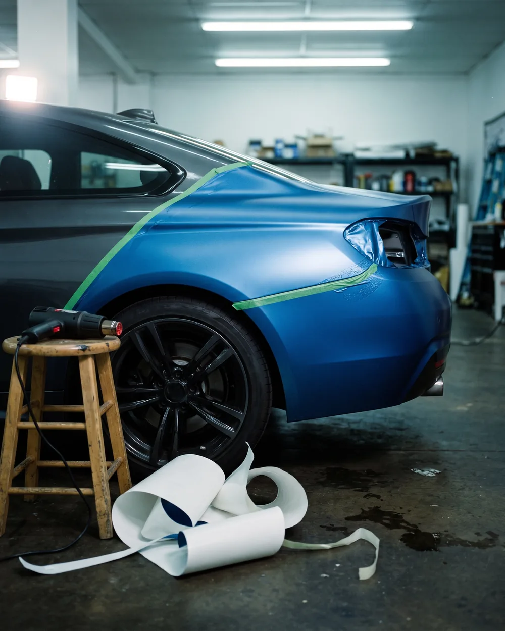 A coupe's rear quarter half wrapped in satin vinyl, a tape line along the seam, a heat gun on a stool.